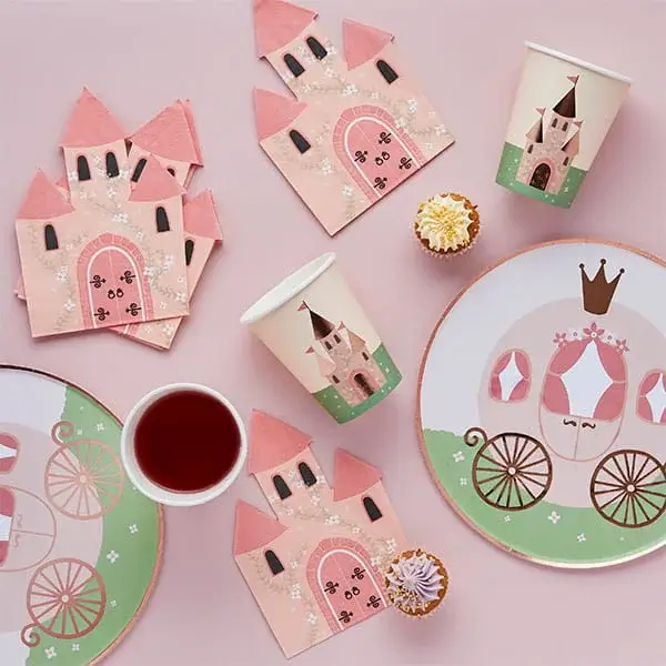 Princess-themed party tableware with castle napkins, carriage plates, cups, and small cupcakes on a pastel pink background.